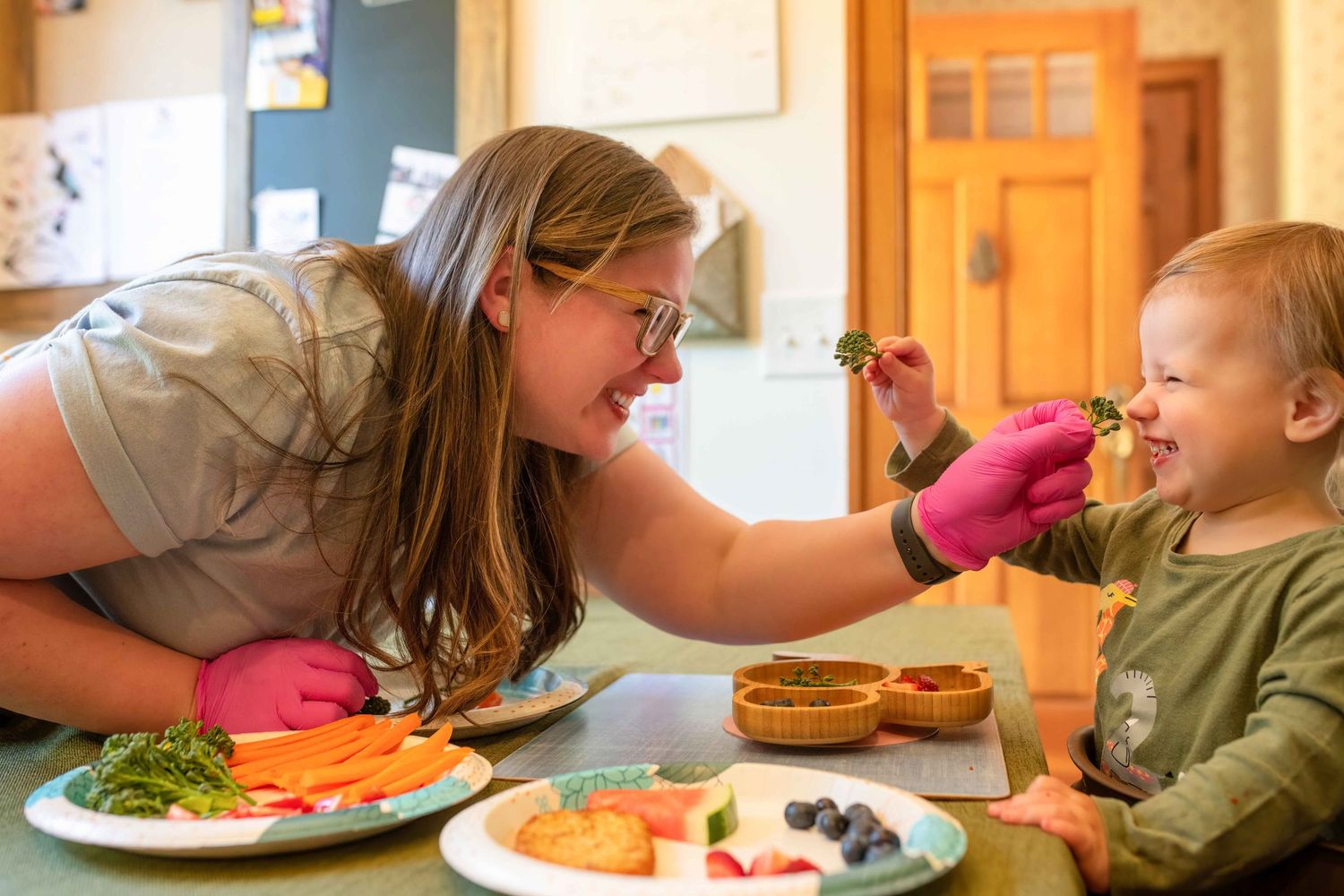 Woman in pink gloves feeds a smiling toddler healthy food at a table with colorful fruits and vegetables.
