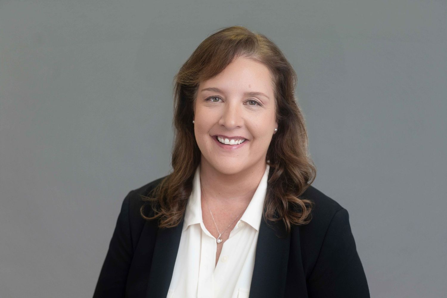 Professional woman in black blazer and white blouse smiling confidently against gray background.