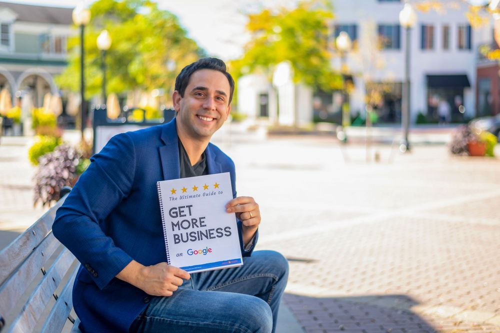Man in blue blazer sitting on bench holding a Get More Business Google sign in an outdoor plaza.