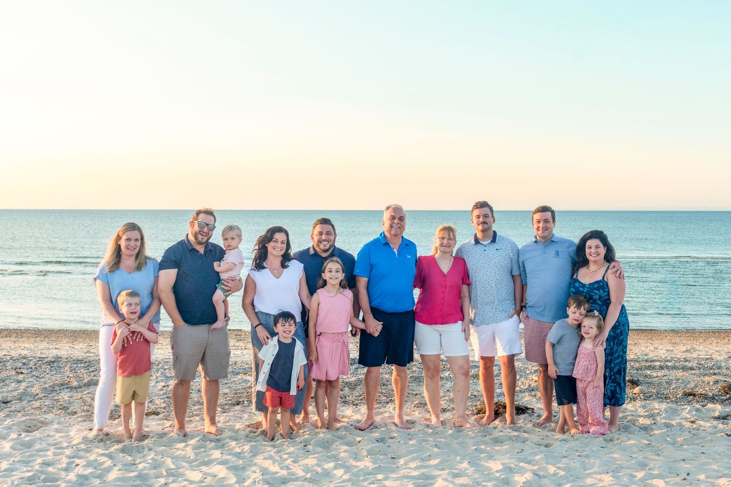 Large family group posing together on a sandy beach at sunset with calm ocean waters in the background.