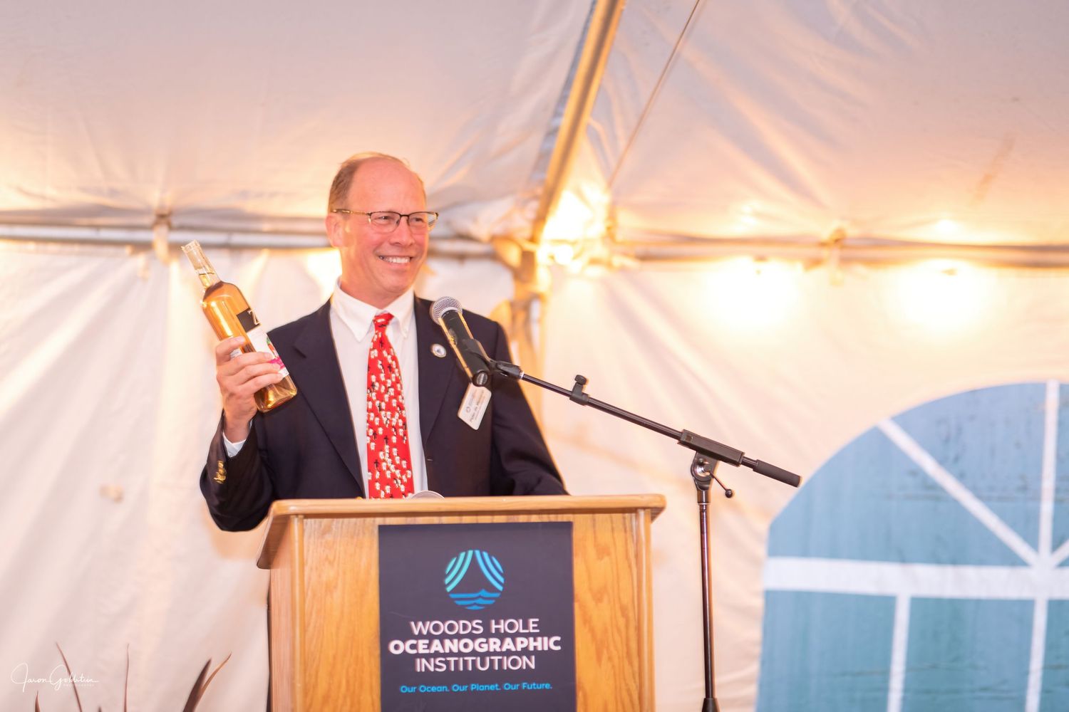 Speaker holding an award while presenting at a podium under event tent lighting.