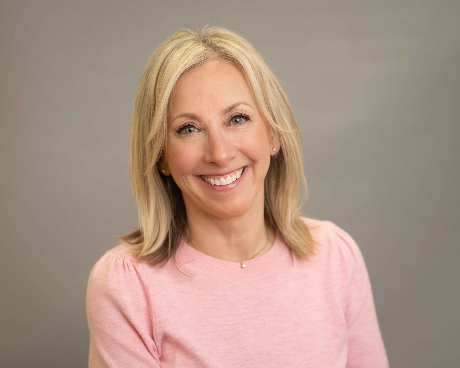 Smiling blonde woman in a pink sweater poses for a professional headshot against a gray background.