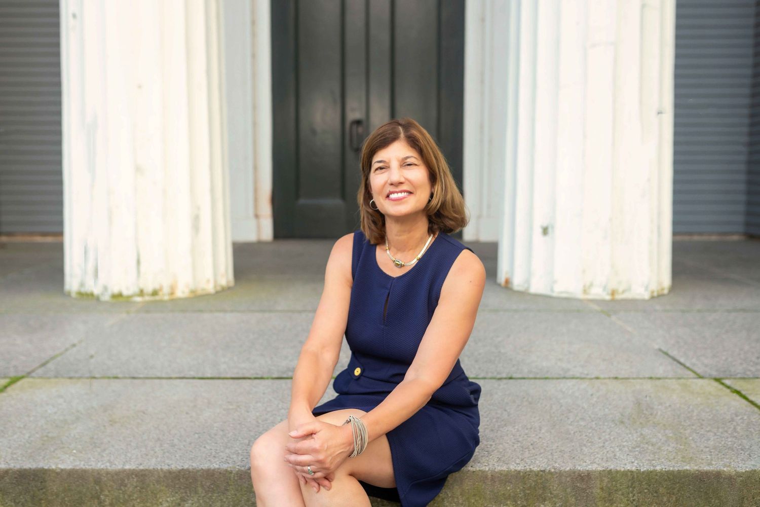 Woman in navy sleeveless dress sitting on steps outdoors, smiling warmly.