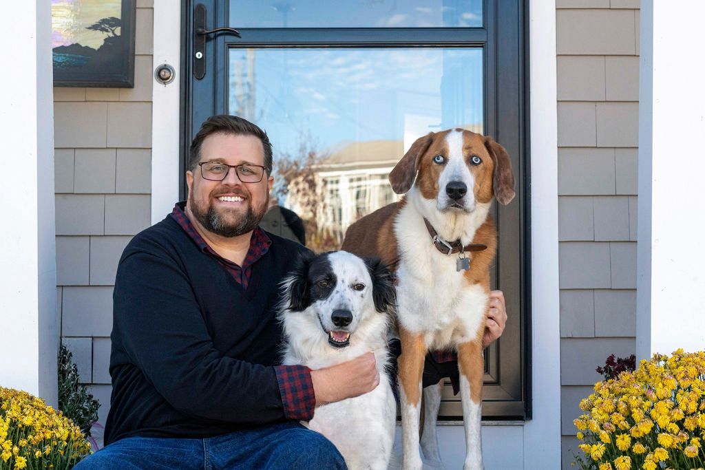 Two dogs and their companion pose cheerfully by front door with yellow flowers.
