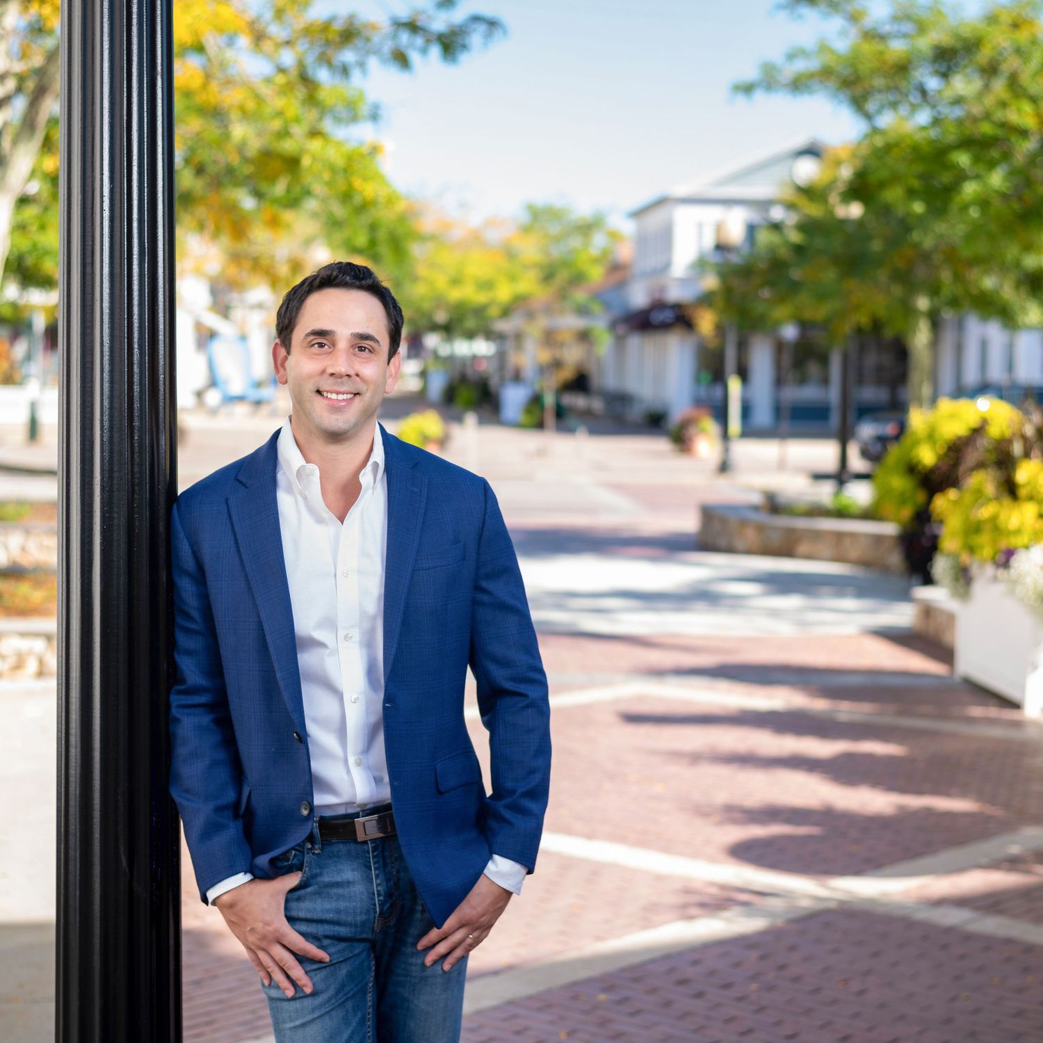 Professional in a navy suit and white shirt stands outdoors on a brick walkway with trees and buildings in background.