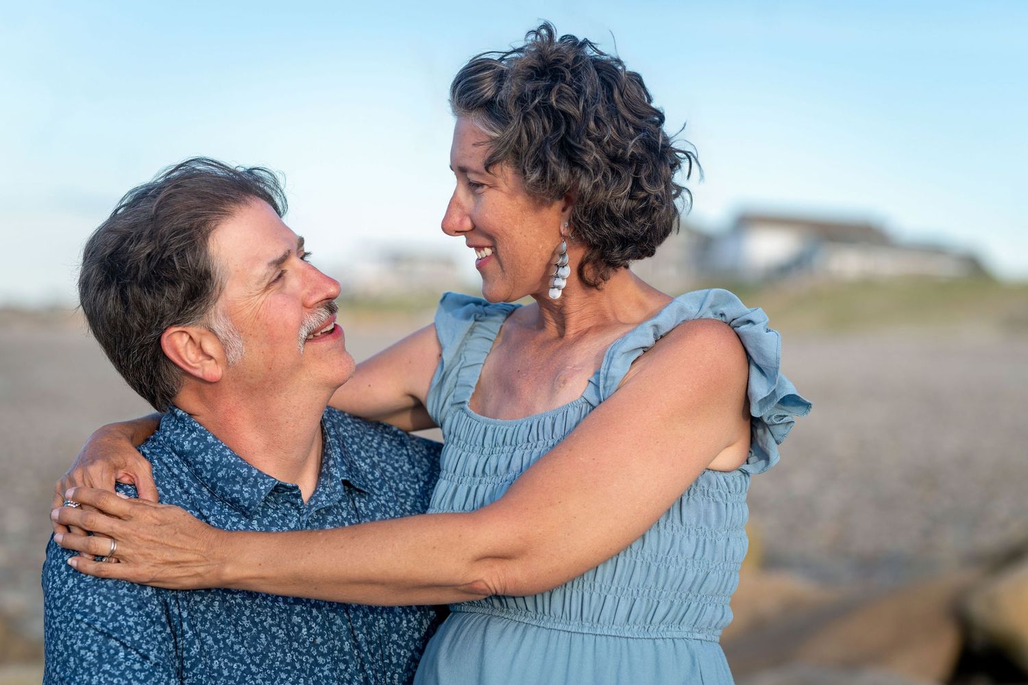 A happy middle-aged couple embracing and smiling at each other outdoors near a beach on a clear day.