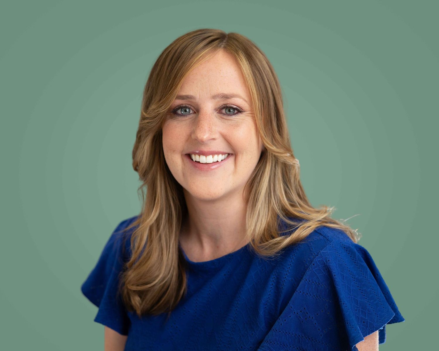 Smiling woman with blonde hair wearing blue top against green background.