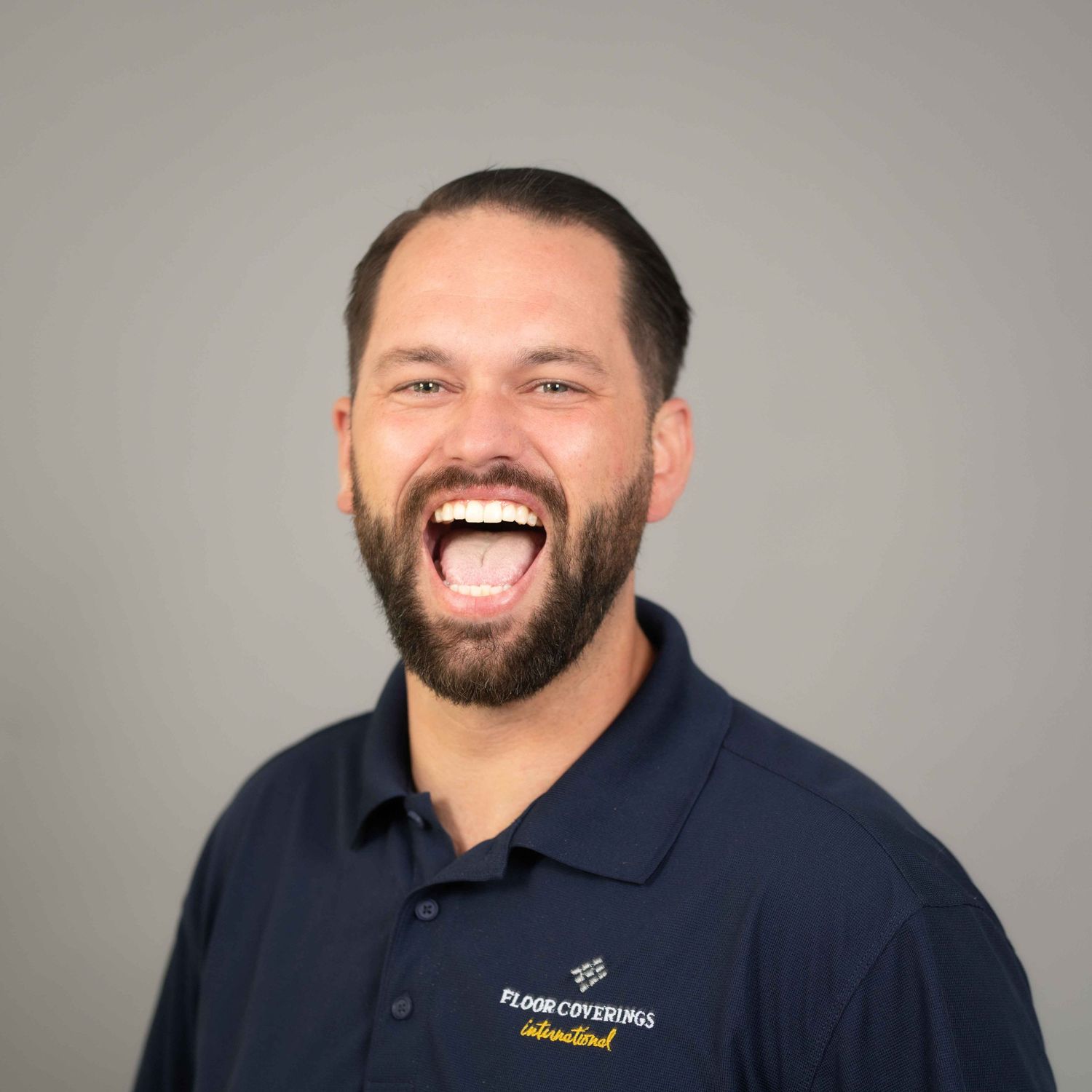 Smiling man with beard wearing a navy Floor Coverings International polo shirt against a gray background.
