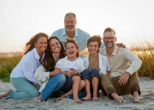 A group poses together on a sandy beach during a sunset photo session in coordinated white and blue outfits.