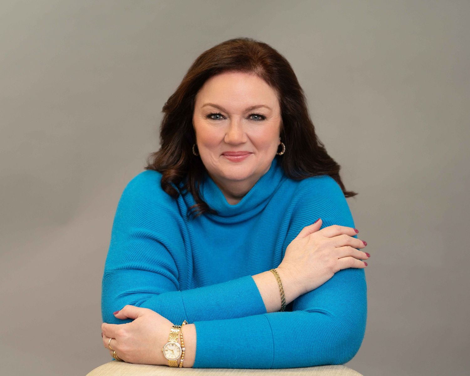 Professional woman in teal turtleneck sweater smiling confidently, wearing gold watch, arms crossed, gray background.