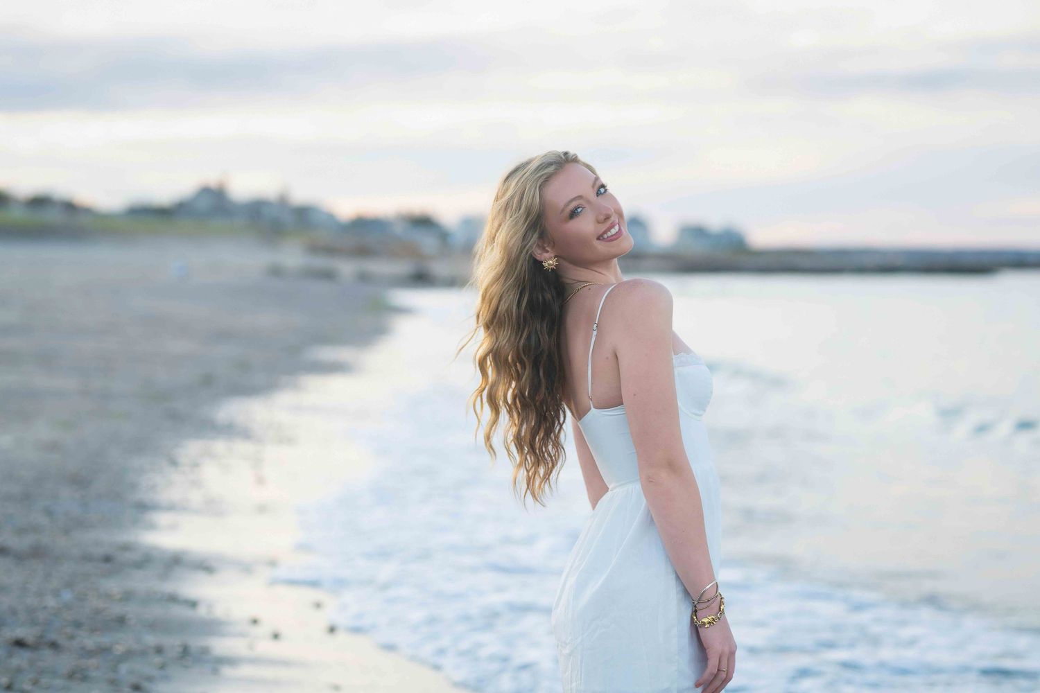 Smiling young woman in white dress standing on a sandy beach with ocean waves and coastal town in background.