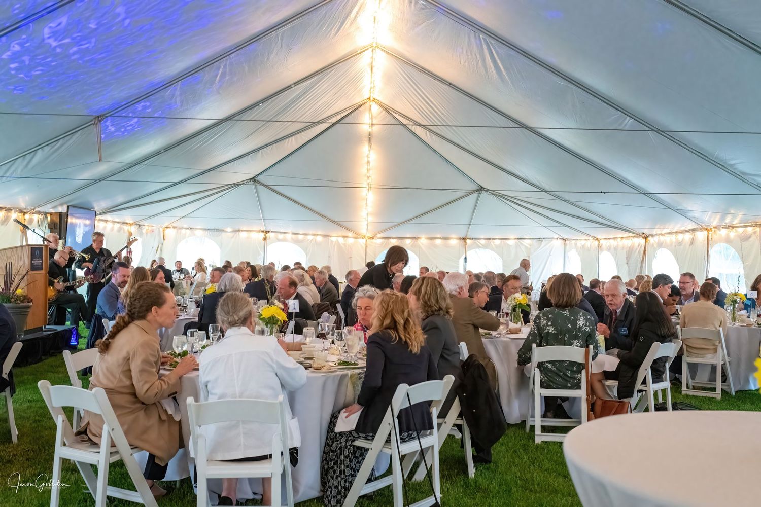 Large white event tent with string lights illuminating the ceiling and guests seated at round tables.