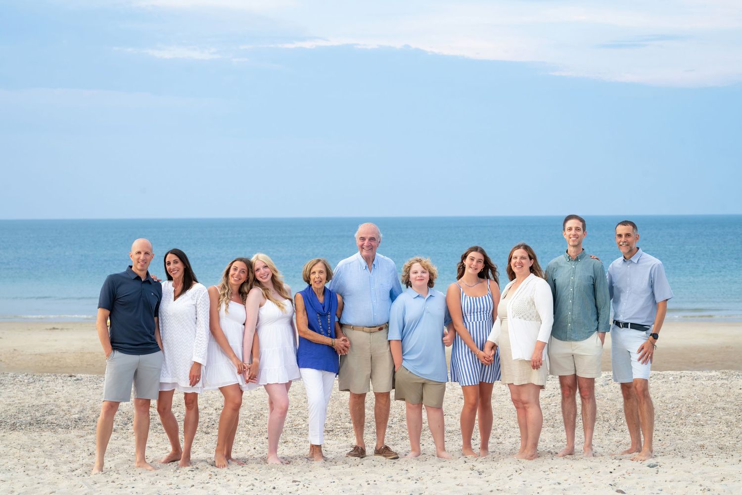 Large family group wearing coordinated white and blue outfits posing on sunny beach.