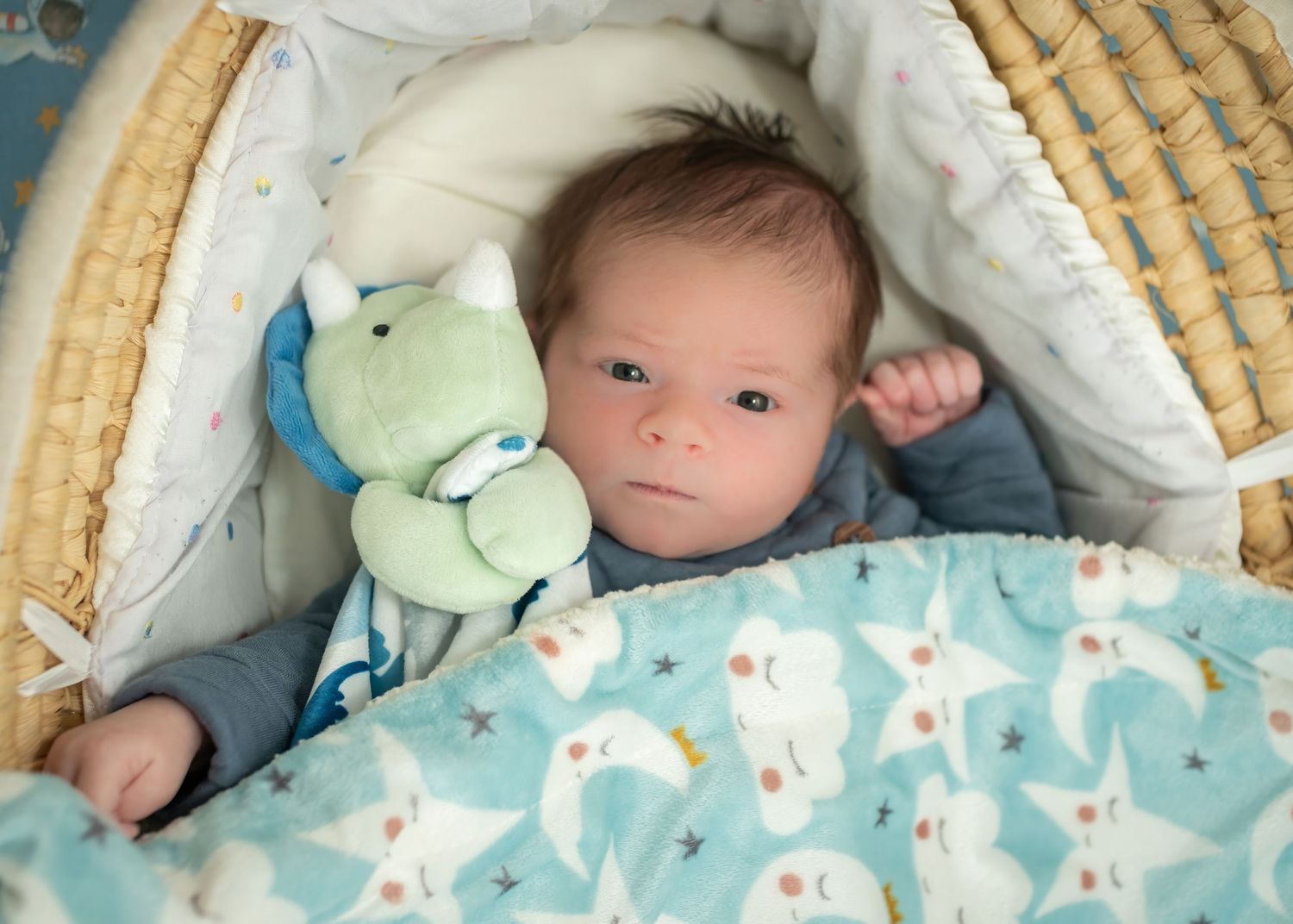Baby resting in bassinet with light blue blanket and plush toys.