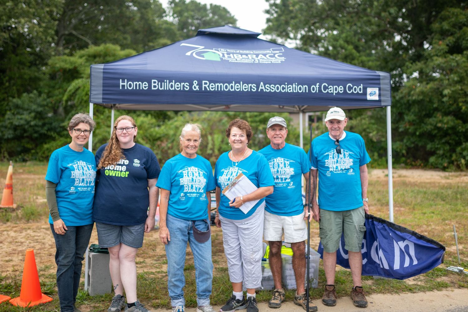 Group of volunteers in blue t-shirts gathered at outdoor community event with tents and banners.