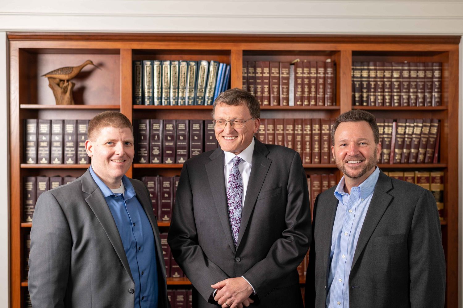 Three professional men in business attire smile in a law office with bookshelves filled with legal volumes.