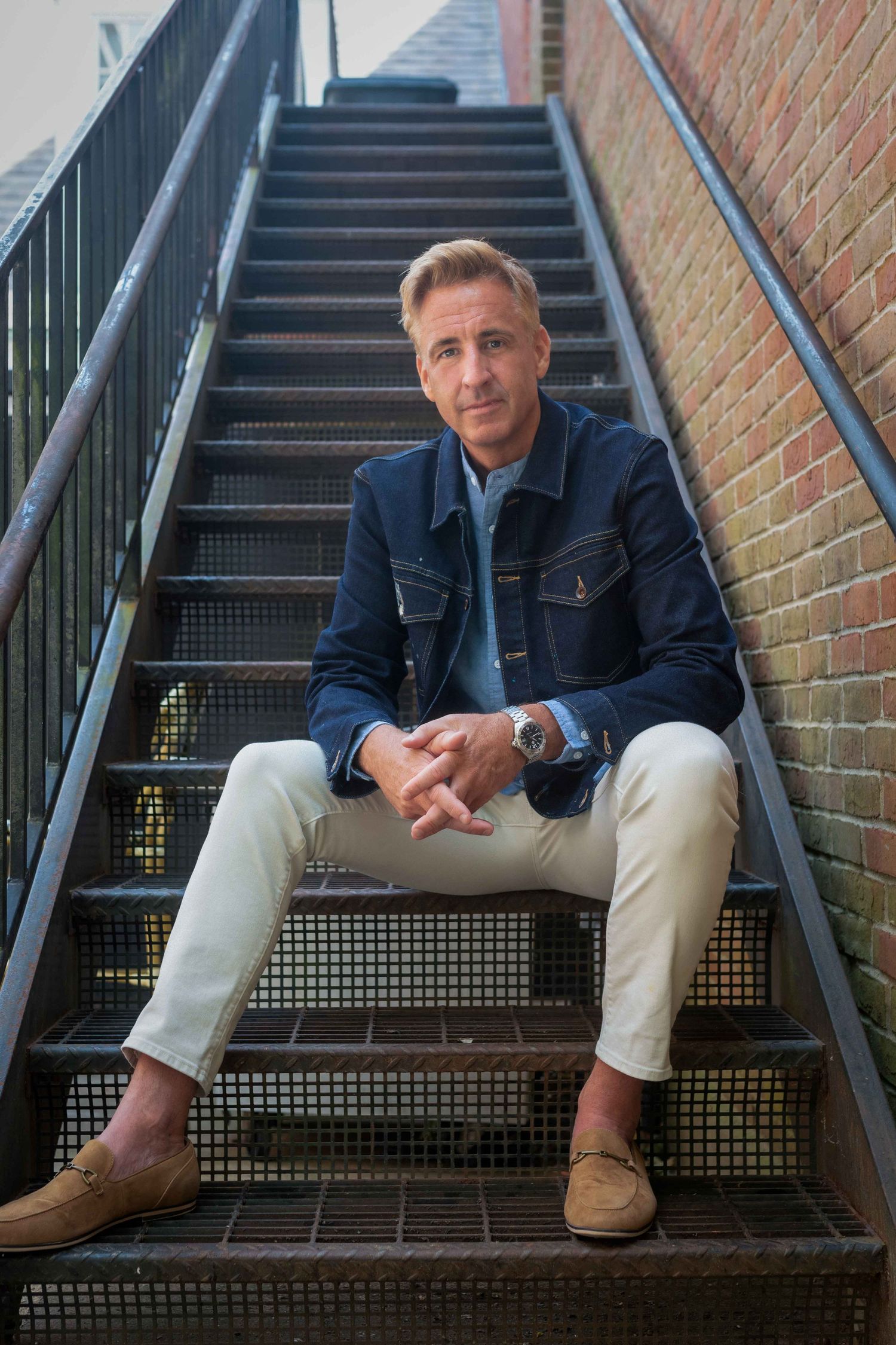 Man in navy jacket and white pants sitting on metal stairs against brick wall.