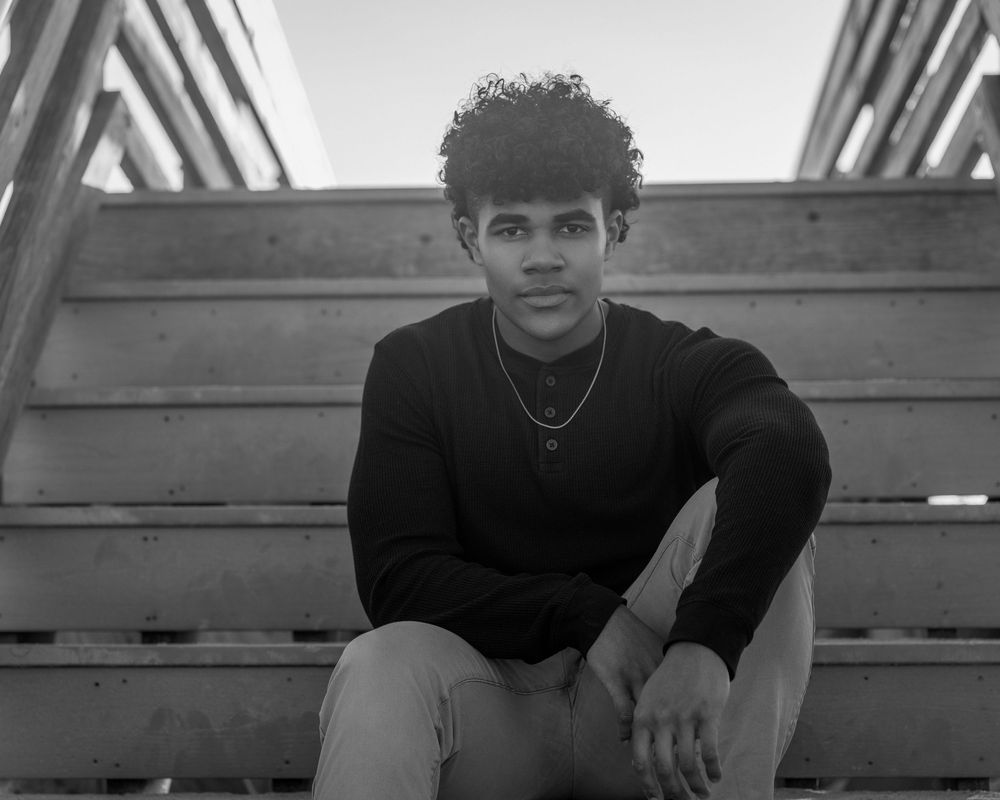 Young man with curly hair sitting on wooden steps, wearing a black sweater, in a black and white photo.