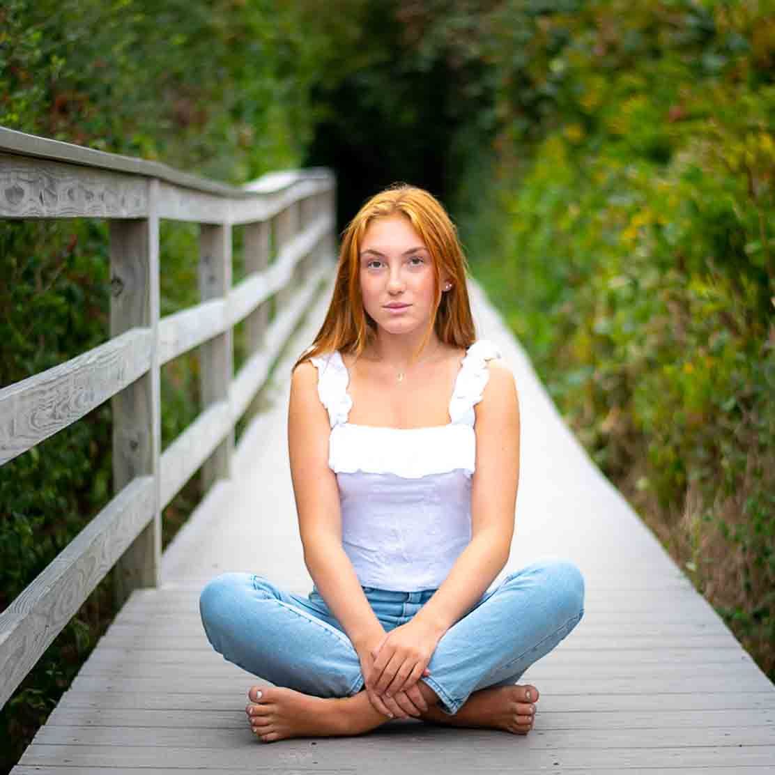 Young woman with red hair sits cross-legged on a wooden bridge path surrounded by lush green foliage.