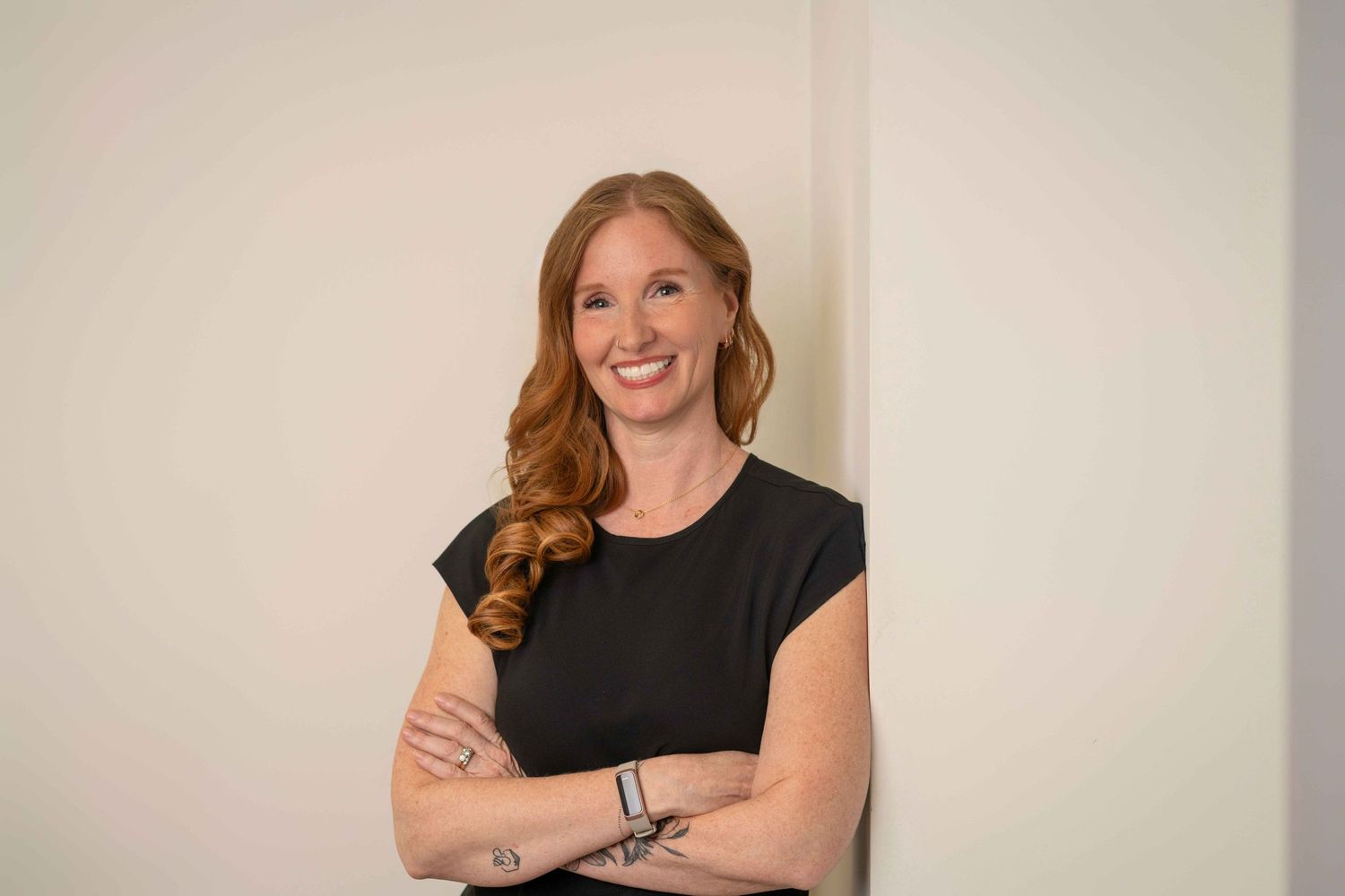 Smiling redhead woman in black top with arms crossed, posing confidently against a white wall.