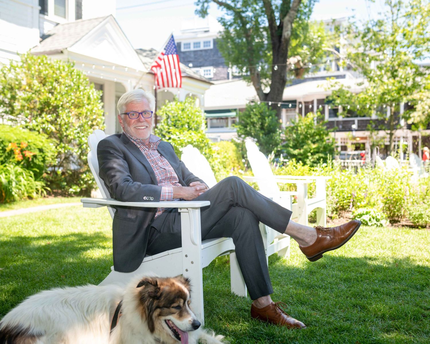 Man in blazer relaxing in white Adirondack chair on green lawn with dog and American flag backdrop.