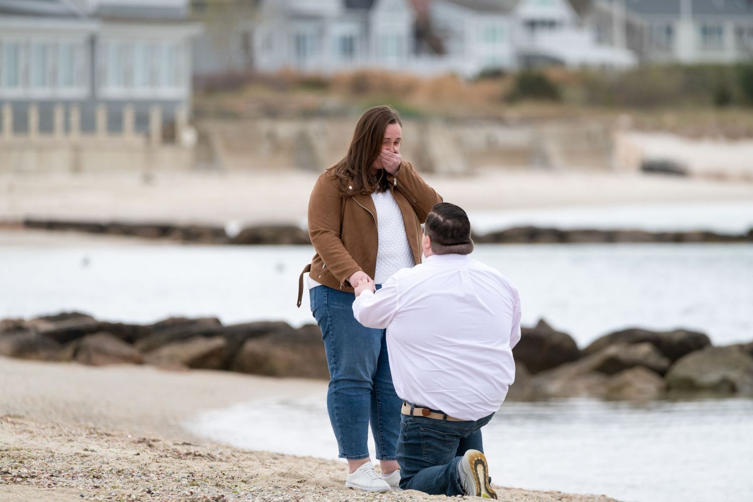 Couple walking along a rocky beach shoreline with houses in the background.