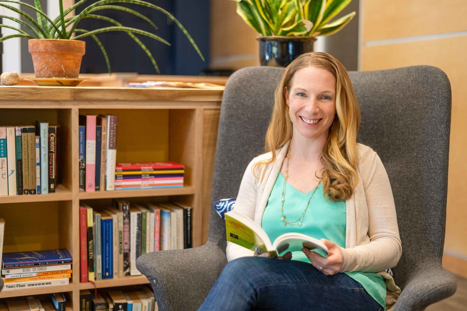 Smiling woman sitting in gray armchair reading a book near a wooden bookshelf with plants in a cozy room.
