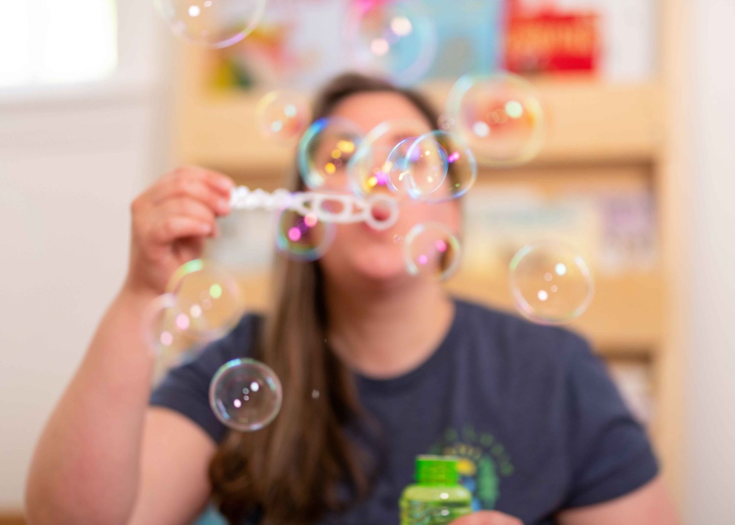 Woman in blue shirt blowing colorful soap bubbles indoors with a bubble wand.