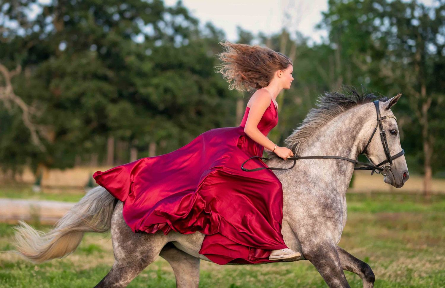 Woman in red gown riding a dapple gray horse through a lush green field, hair flowing in the wind.
