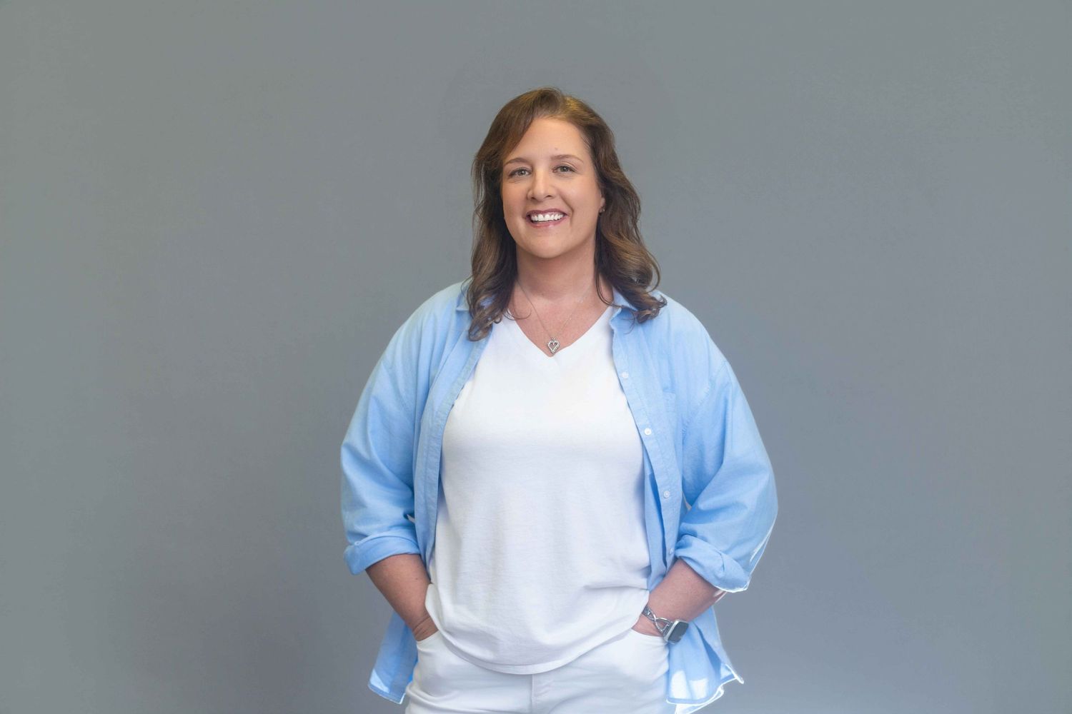 Smiling woman in blue shirt and white top posing confidently against gray background.