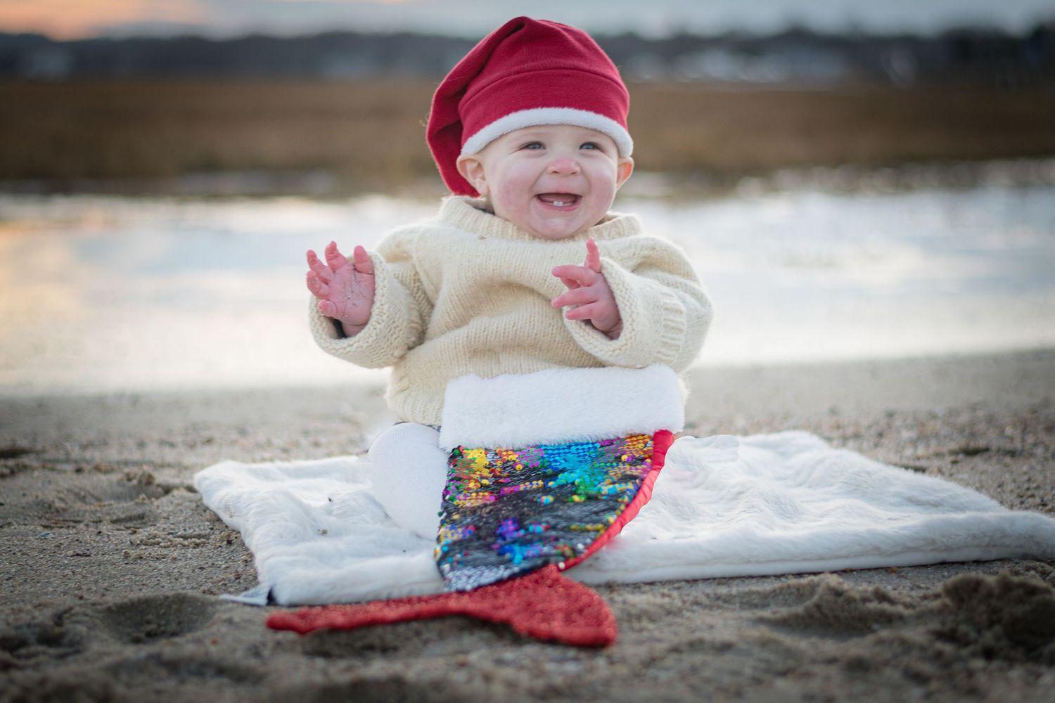 Baby lying on white blanket in snow wearing red winter hat.