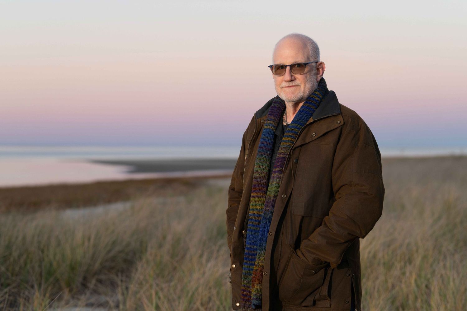 Bald man in sunglasses wearing a colorful scarf and dark coat stands on a sandy beach at sunset.