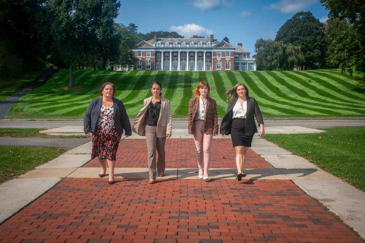 Four professional women walking on a brick path in front of a grand estate with manicured green lawns.