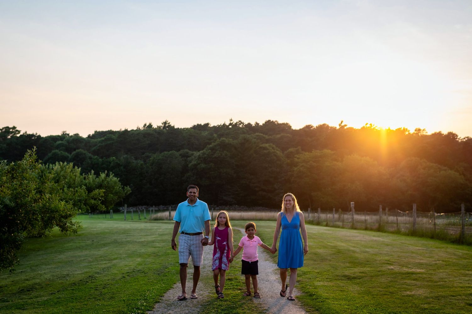 Family walking together on a grassy path at sunset with trees silhouetted against the orange sky.