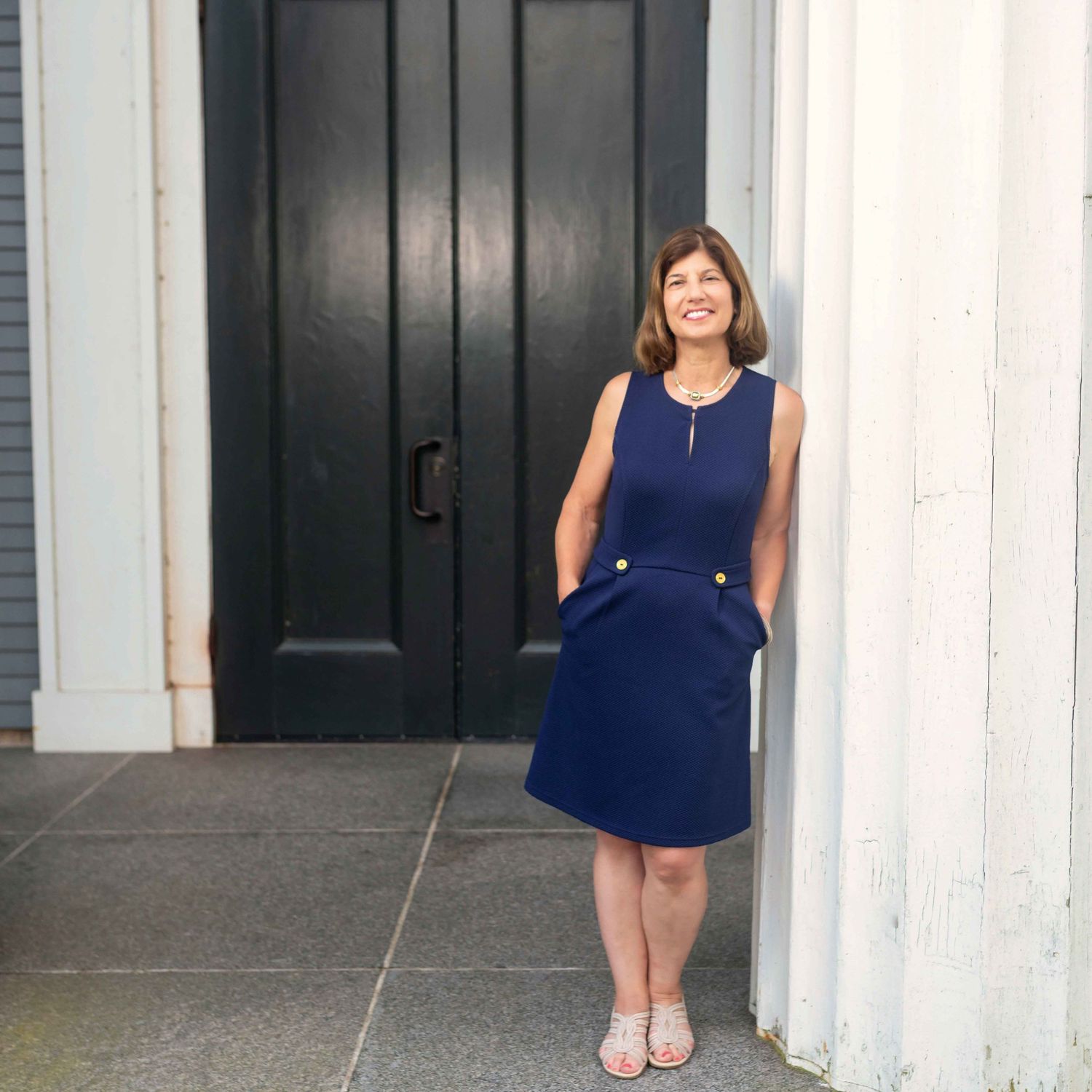 Woman in navy blue sleeveless dress smiling beside a black door and white column outdoors.