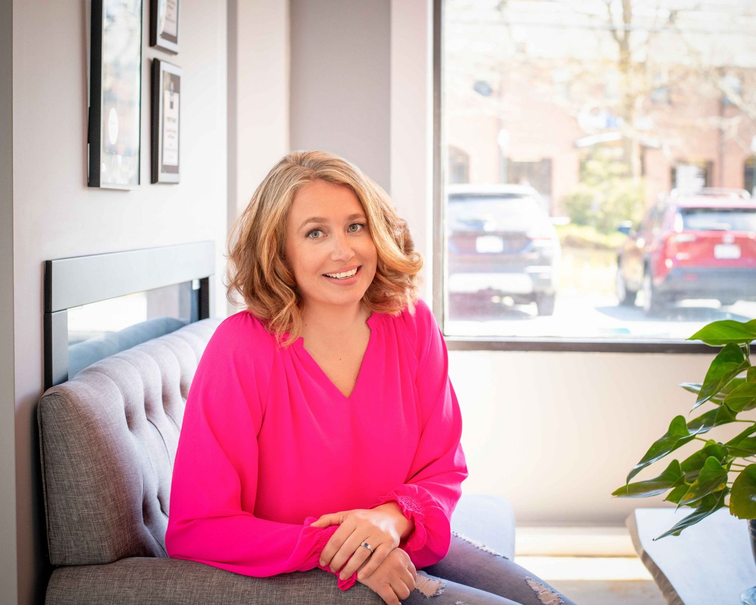 Smiling blonde woman in a bright pink blouse sitting on a gray sofa near a window in a modern office.