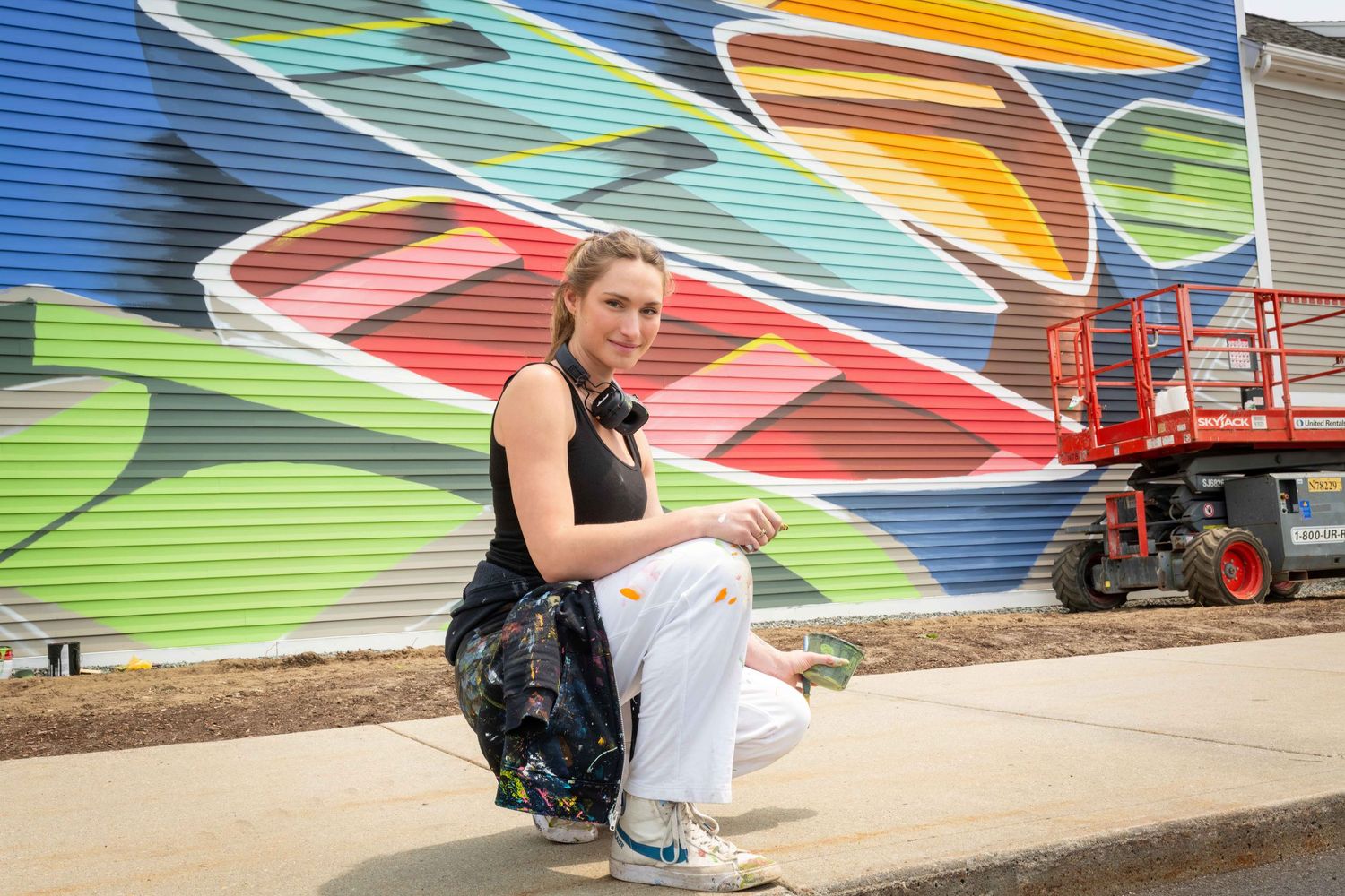 Artist crouching in front of a vibrant colorful mural on a building exterior on a sunny day.