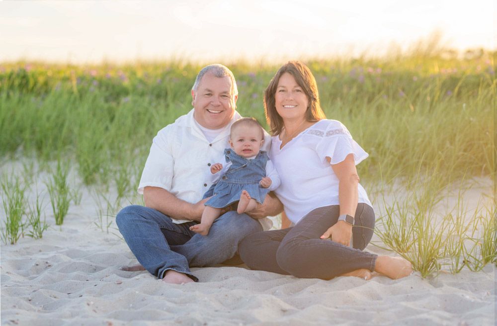 Happy family of three sitting on sandy beach with dunes, parents smiling with baby in blue outfit at golden hour.