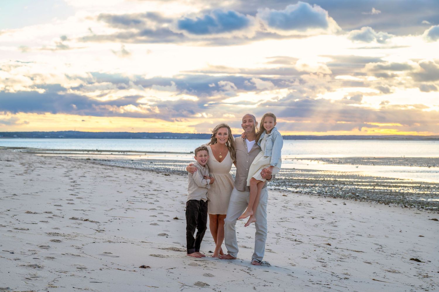 A group stands together on a sandy beach at sunset with dramatic clouds and ocean waves in the background.