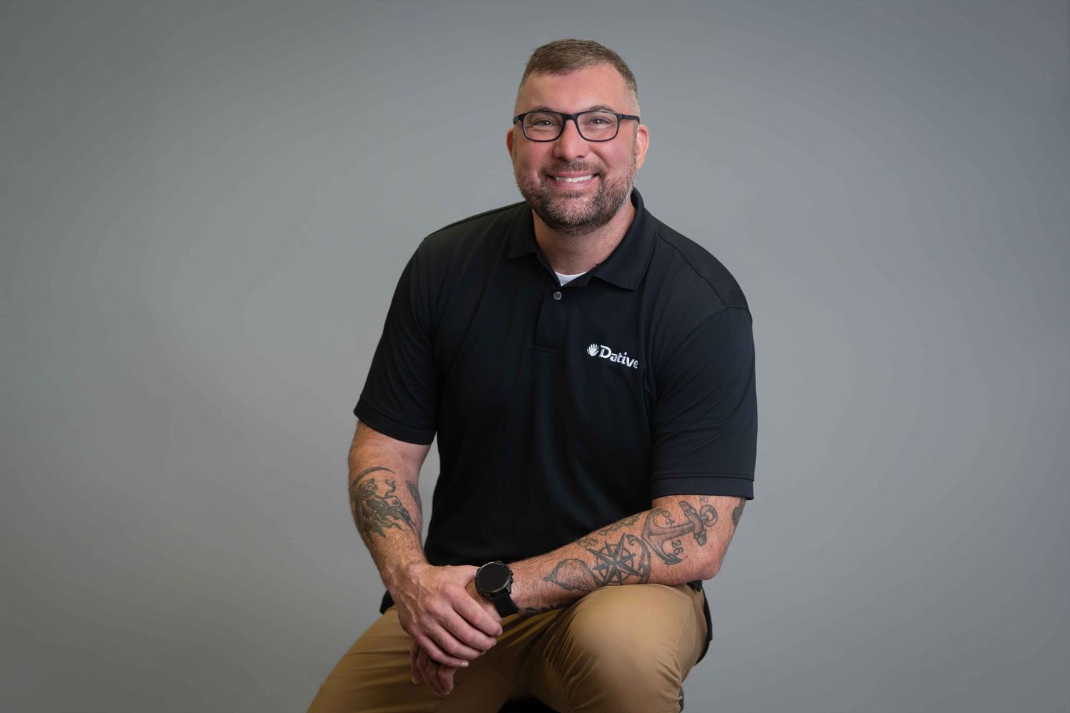 Tattooed man in black polo shirt sitting casually and smiling against a gray background.