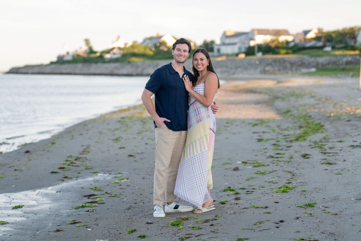 A couple takes a romantic stroll along a beach with boats visible in the harbor.