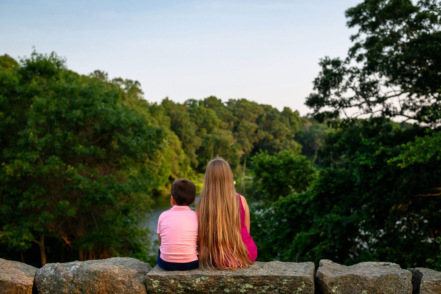 Two people sit together on a stone wall overlooking a scenic wooded area at sunset.