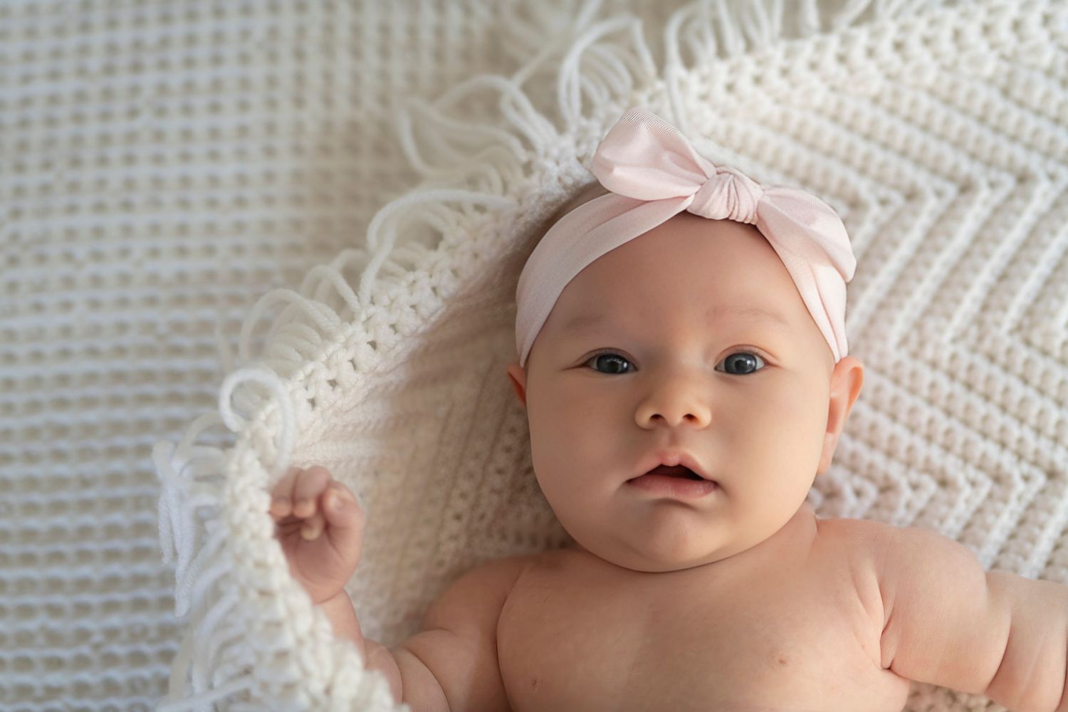 Baby wearing pink headband lying on white knit blanket.