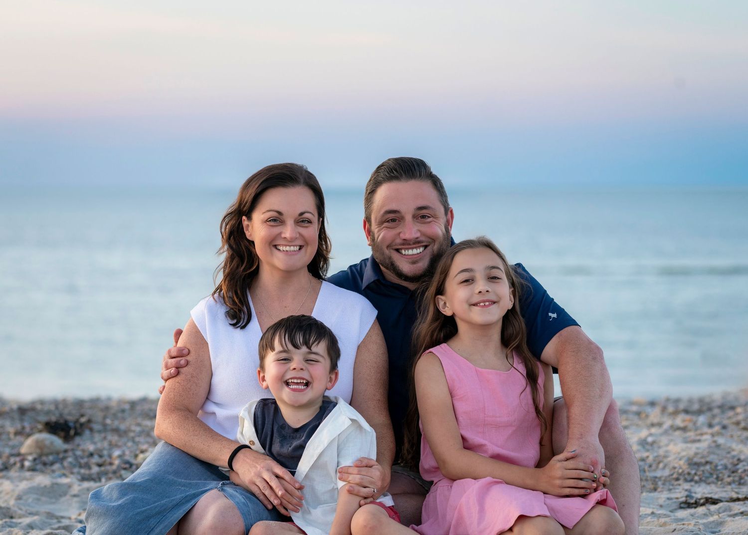 Happy family of four smiling together on a rocky beach at sunset, parents with young boy and girl in pink dress.