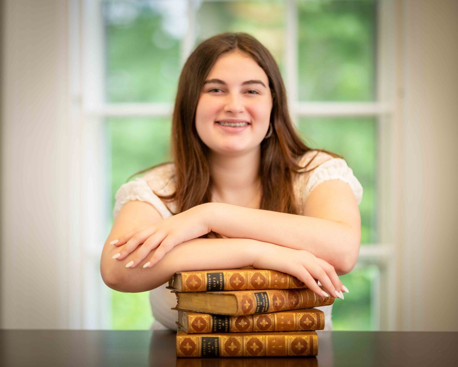 Smiling teenage girl resting arms on stack of vintage books at a table near a bright window.