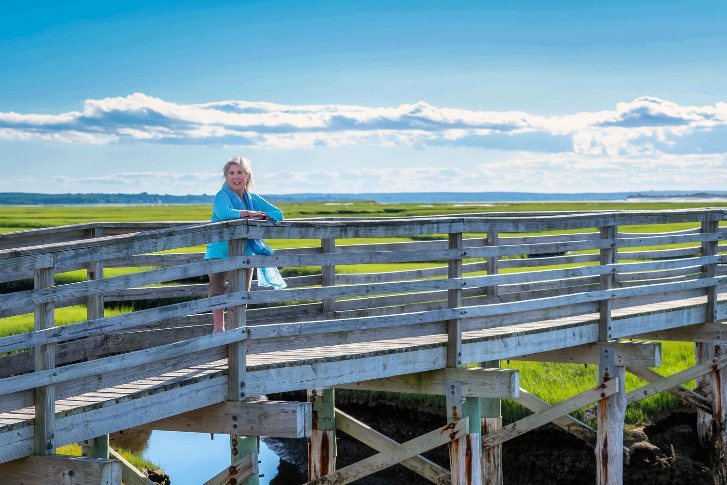 Woman in blue jacket standing on wooden boardwalk bridge overlooking green marshlands under blue sky with clouds.