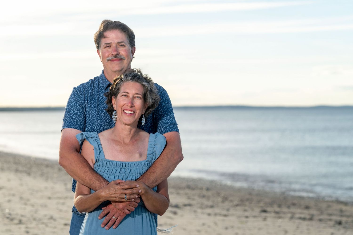Happy couple embracing on a sandy beach at sunset, smiling warmly with calm ocean waters in the background.
