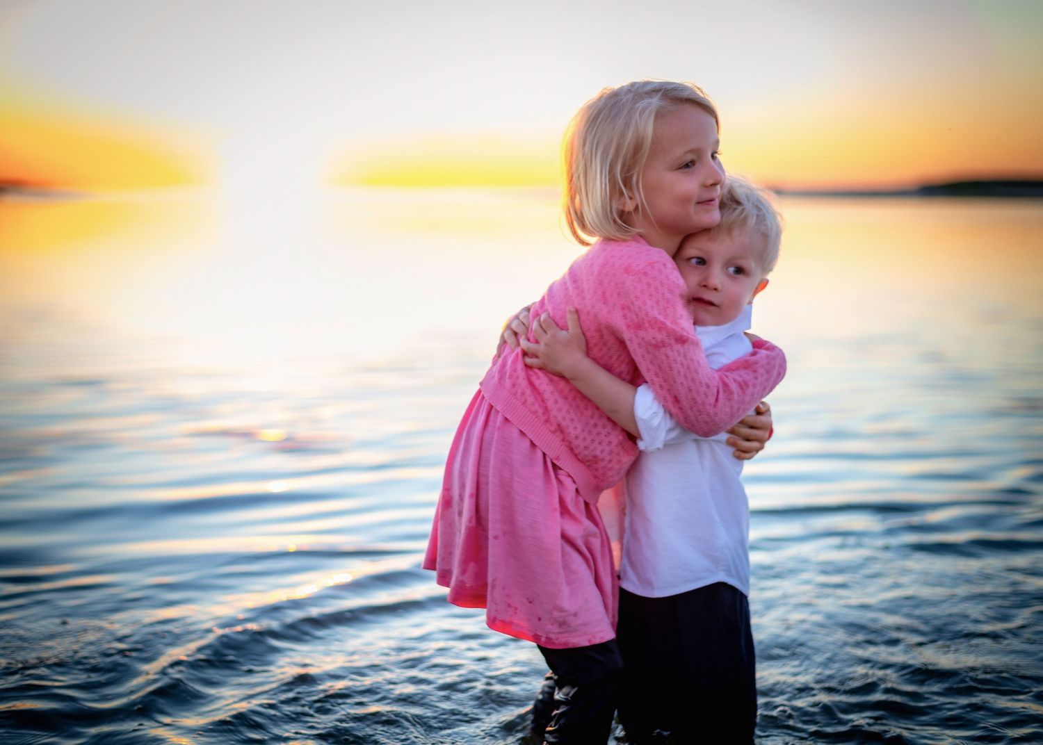Children hugging in pink outfits against sunset ocean backdrop.