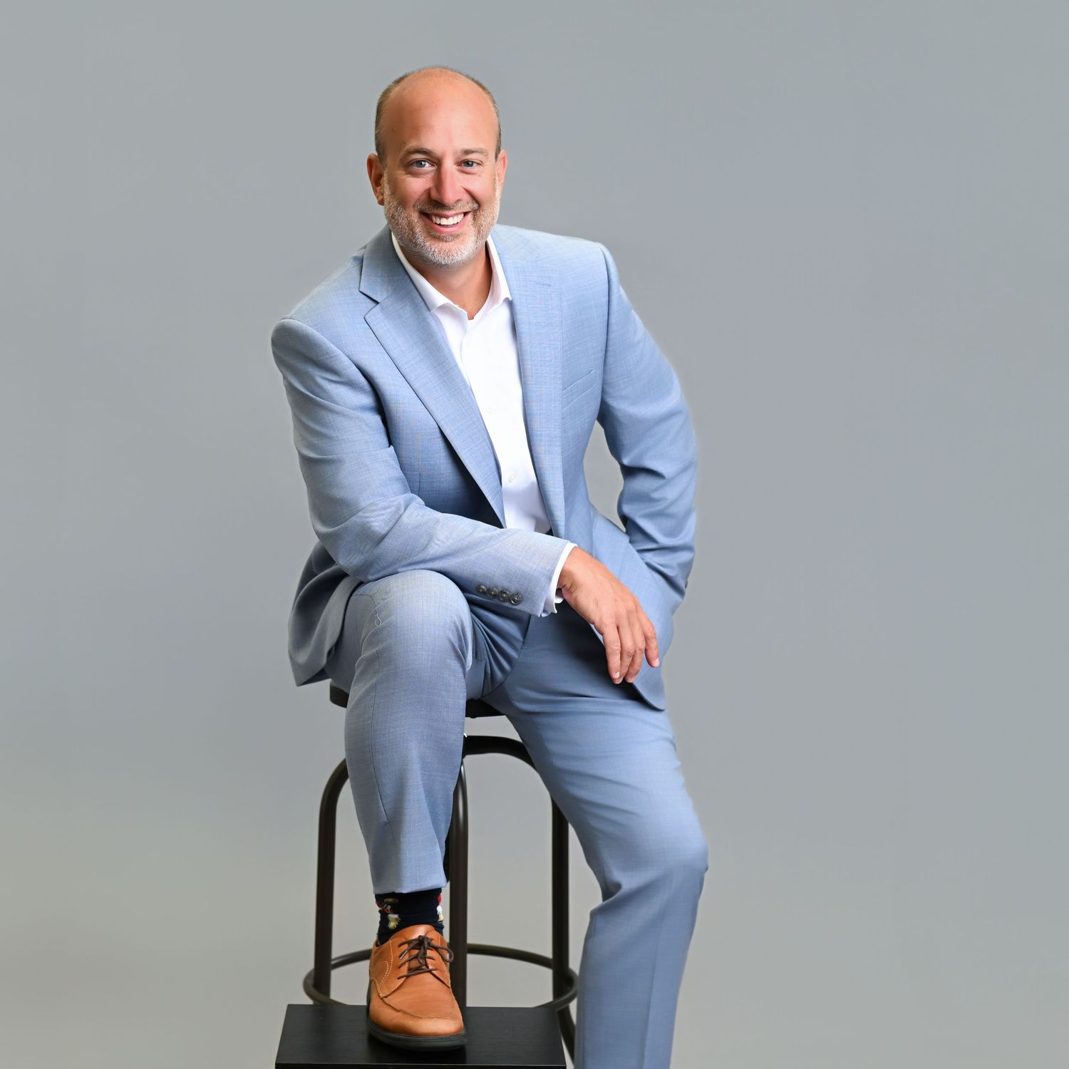 Professional in light blue suit seated on stool against gray backdrop in four different casual poses.