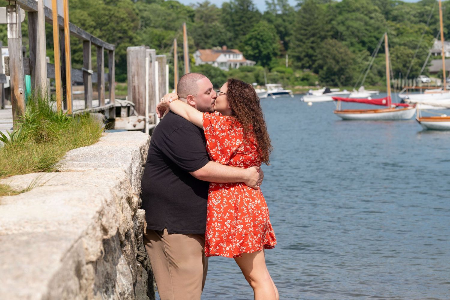 Couple embracing near waterfront with boats and harbor in background.
